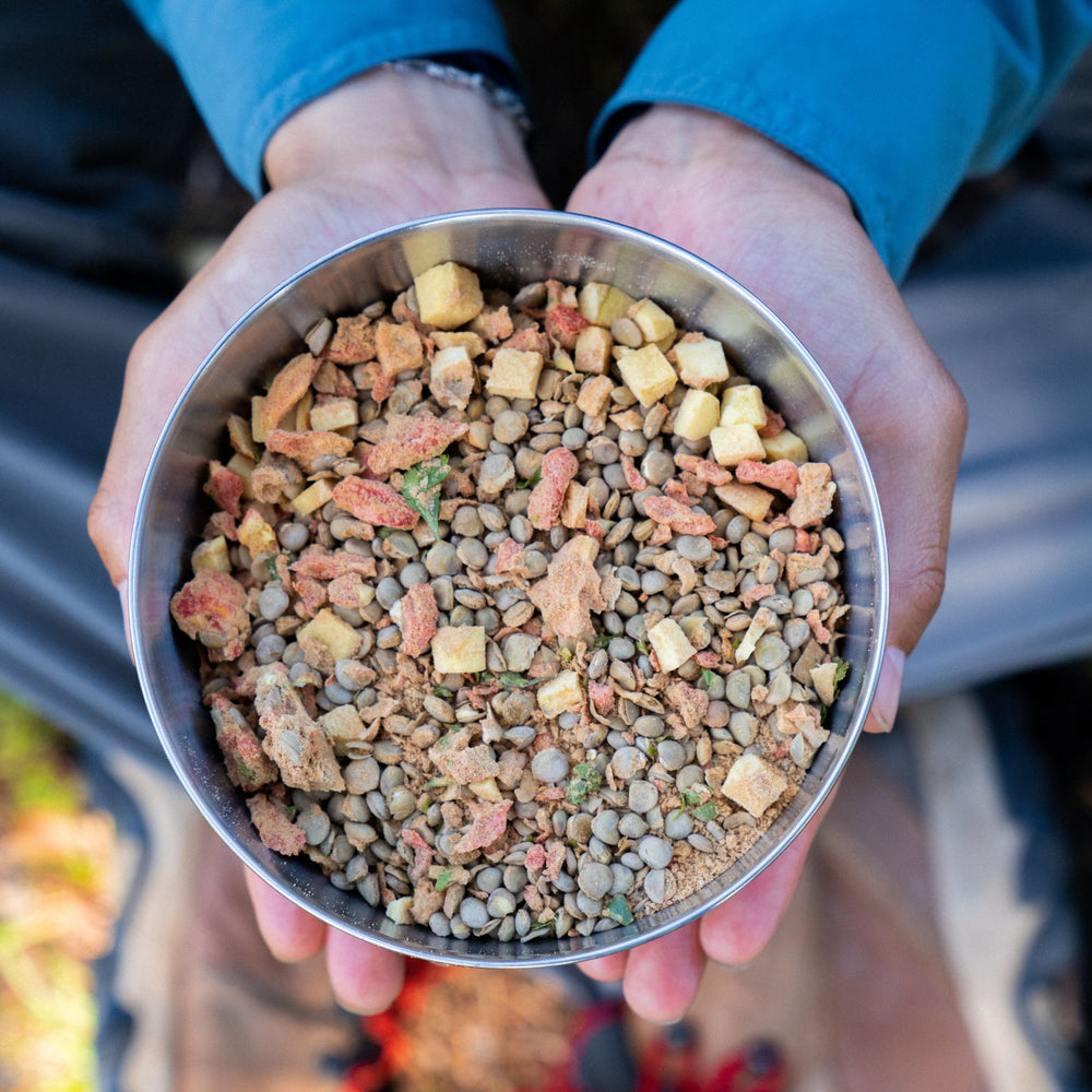 A birdseye view of two hands holding a silver outdoor bowl filled with dehydrated Itacate Campsite Lentejas 