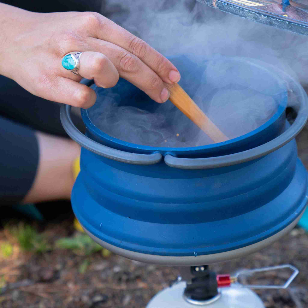 a hand stirring arroz con leche in a pot using a canister camp stove